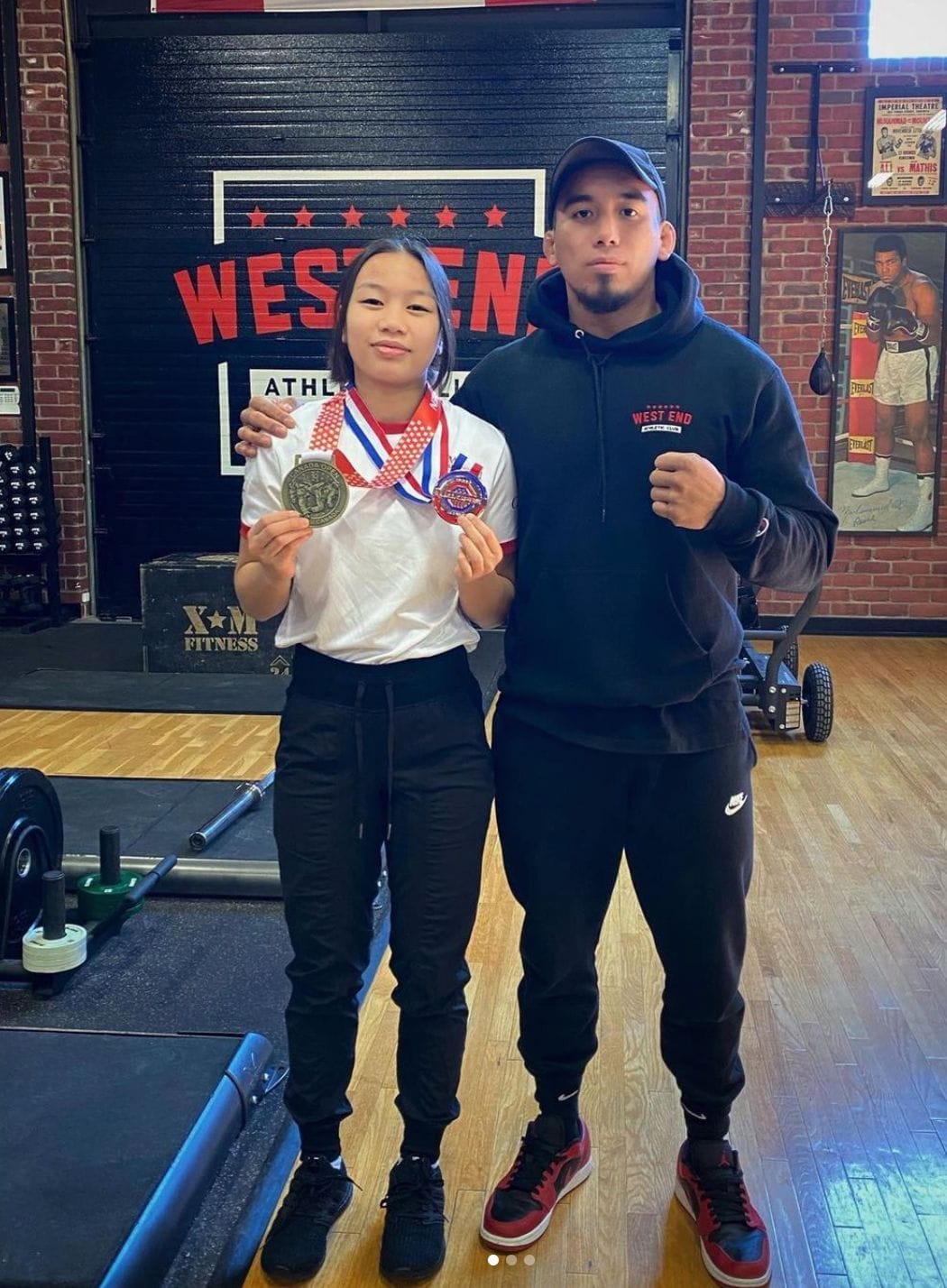 Isla standing beside Coach Miguel inside West End AC holding her gold medal from the Canada Open in Vancouver and her bronze medal from the US Open in Las Vegas