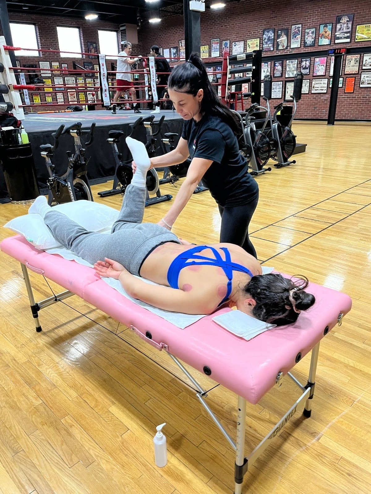  Melissa Karagianis, West End Athletic Club&rsquo;s Certified Manual Osteopath, is standing beside a pink table with a client lying face down. The table is set outside of the boxing ring at West End AC, cupping marks are visible on the client's upper back, and Melissa is holding up the client's left leg at a 90-degree angle.