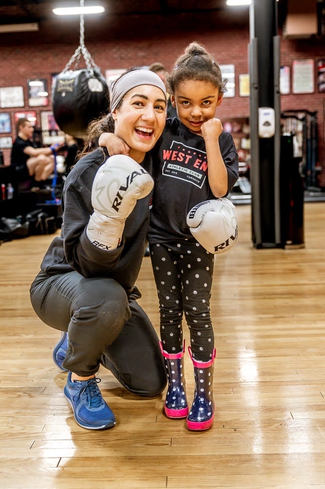 West End Athletic Club boxing coach Sara Haghighat-joo wearing white boxing gloves with her arm around a young girl wearing a black t-shirt in the West End AC gym in Toronto.