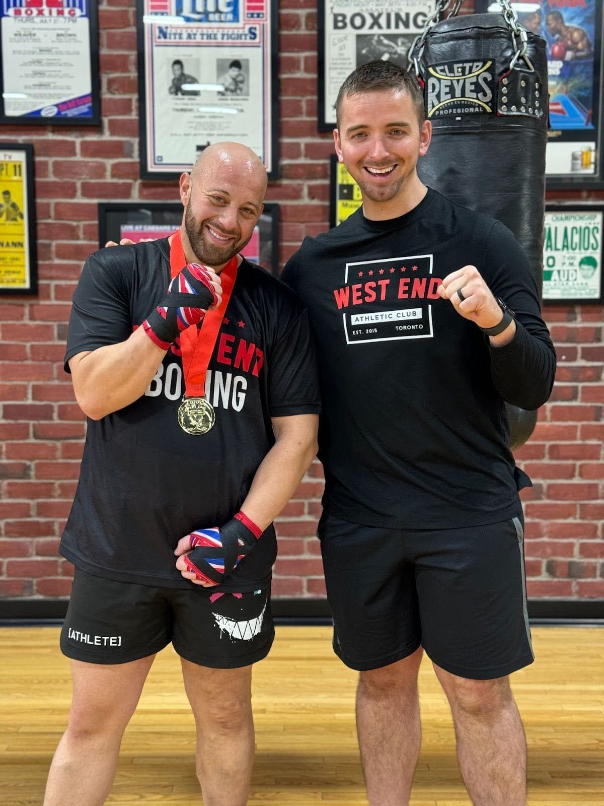 Coach Stevie Bailey and Danny Quattrociocchi both smiling, standing beside each other, in front of a punching bag at West End Athletic Club in Toronto.