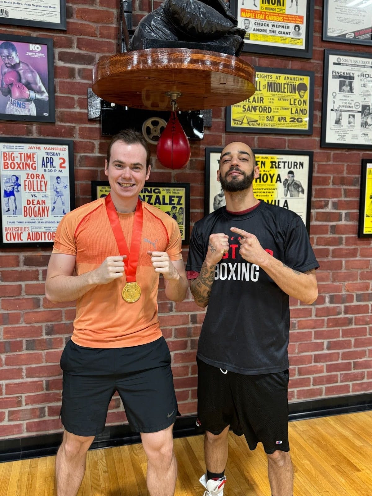 Niles Santos is pointing to his right at member Julian Pettit who is smiling and wearing his medal. They are inside the West End Athletic Club boxing and fitness gym, standing under a red speed bag.