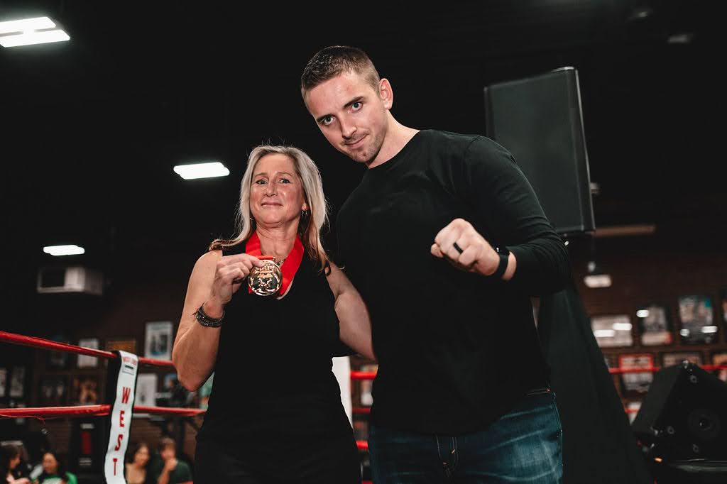 Coach Stevie Bailey is standing inside the boxing ring at Toronto&rsquo;s West End Athletic Club beside Ann Frazer who is wearing all black and holding her medal.